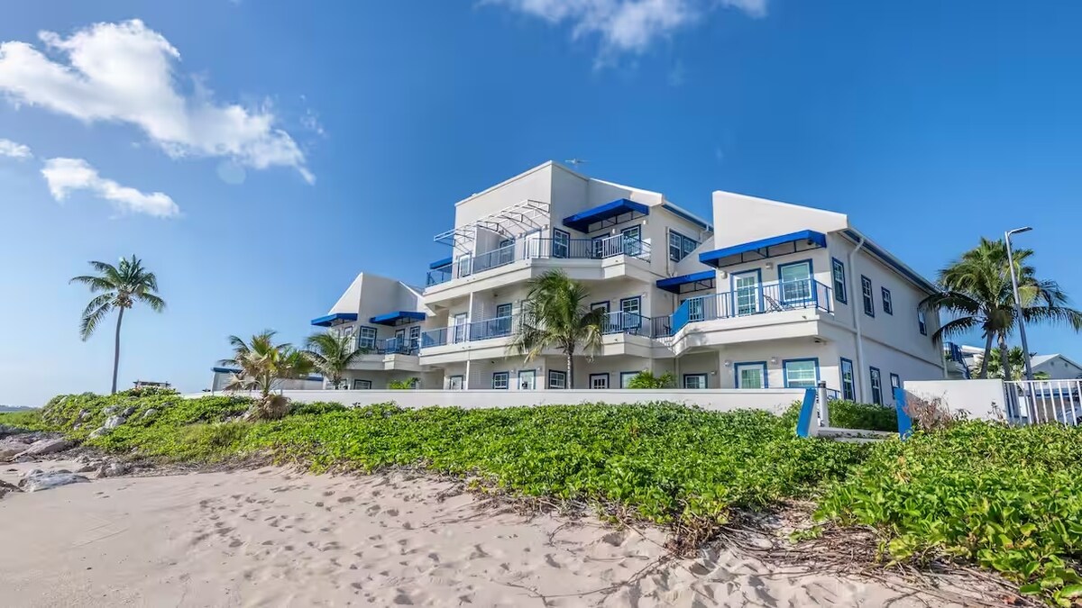 A beachfront resort is visible, featuring a three-story structure with multiple balconies overlooking the ocean. Lush greenery borders the sandy beach, while clear blue skies and a few scattered clouds enhance the coastal setting. Palm trees gently sway in the foreground.