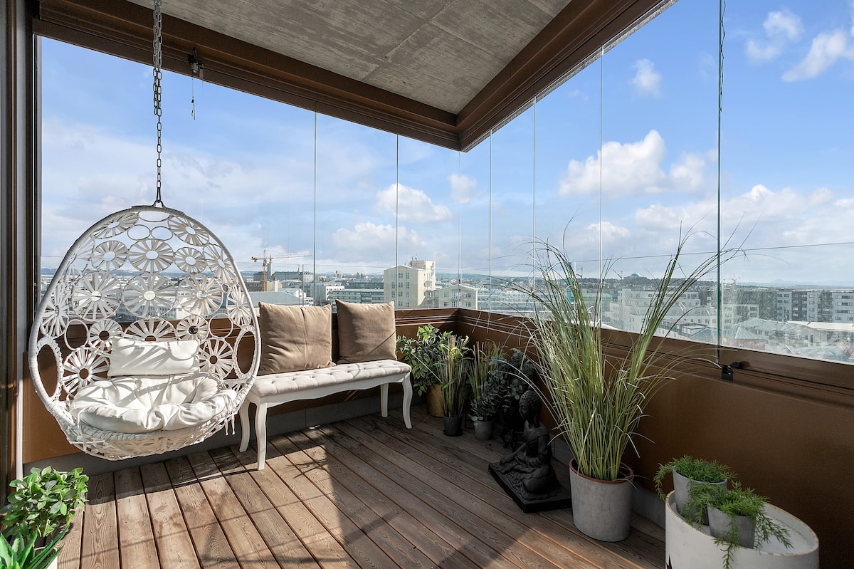 A balcony is framed by expansive glass panels, offering unobstructed views of the surrounding area. A hanging chair with a cushion complements a wooden bench. Potted plants enhance the space, adding greenery against a backdrop of a clear blue sky.
