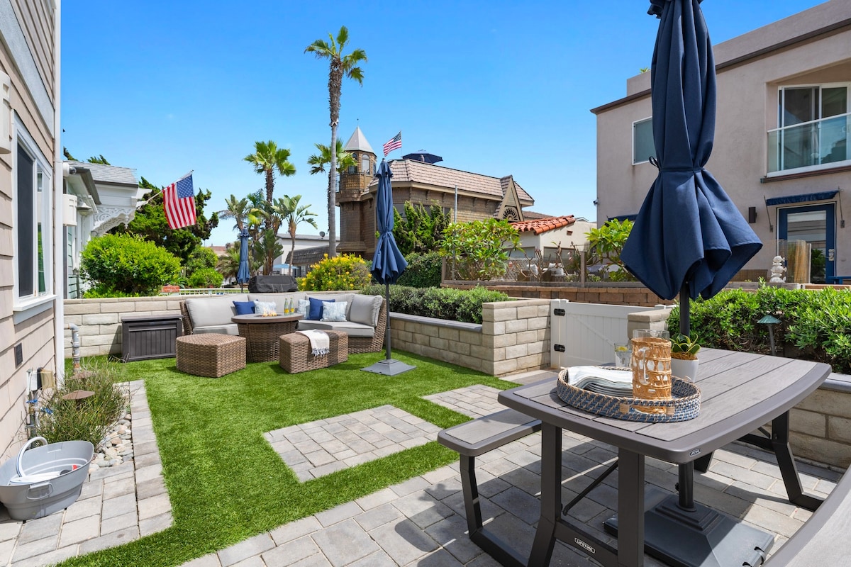 An outdoor patio space features lounge seating arranged around a fire pit, surrounded by greenery. A picnic table with an umbrella is positioned nearby, while the area is paved with stone. Decorative elements include flags and tropical palm trees in the background.