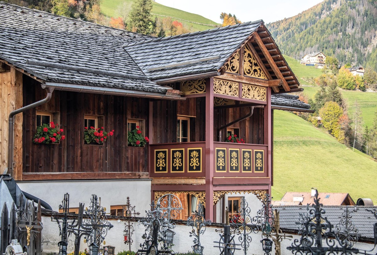 A charming wooden building is depicted with intricate designs and colorful flower boxes adorning the windows. The rustic architecture features a detailed overhang, with lush green hills serving as a backdrop, enhancing the natural setting of the holiday apartment.