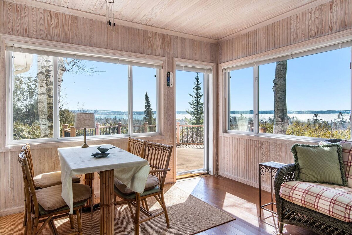 A three-season sunroom is showcased, featuring large windows that offer views of the surrounding trees and water. A dining table with place settings is positioned in the center, complemented by a cozy seating area adorned with cushions. Light wood paneling enhances the tranquil atmosphere.