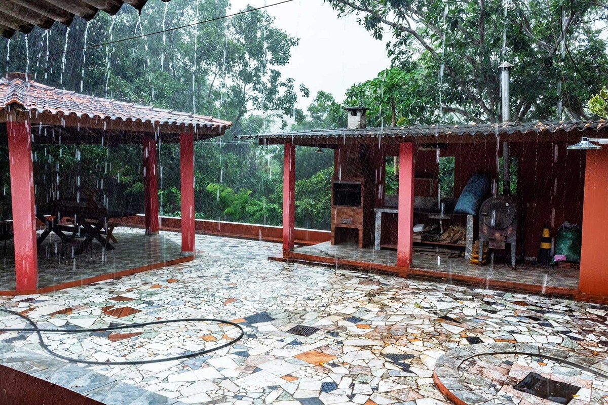 A covered outdoor area is captured amidst rainfall, showcasing a spacious patio with a patterned stone floor and a rustic barbecue station. Lush greenery is visible in the background, enhancing the sense of nature surrounding the space.
