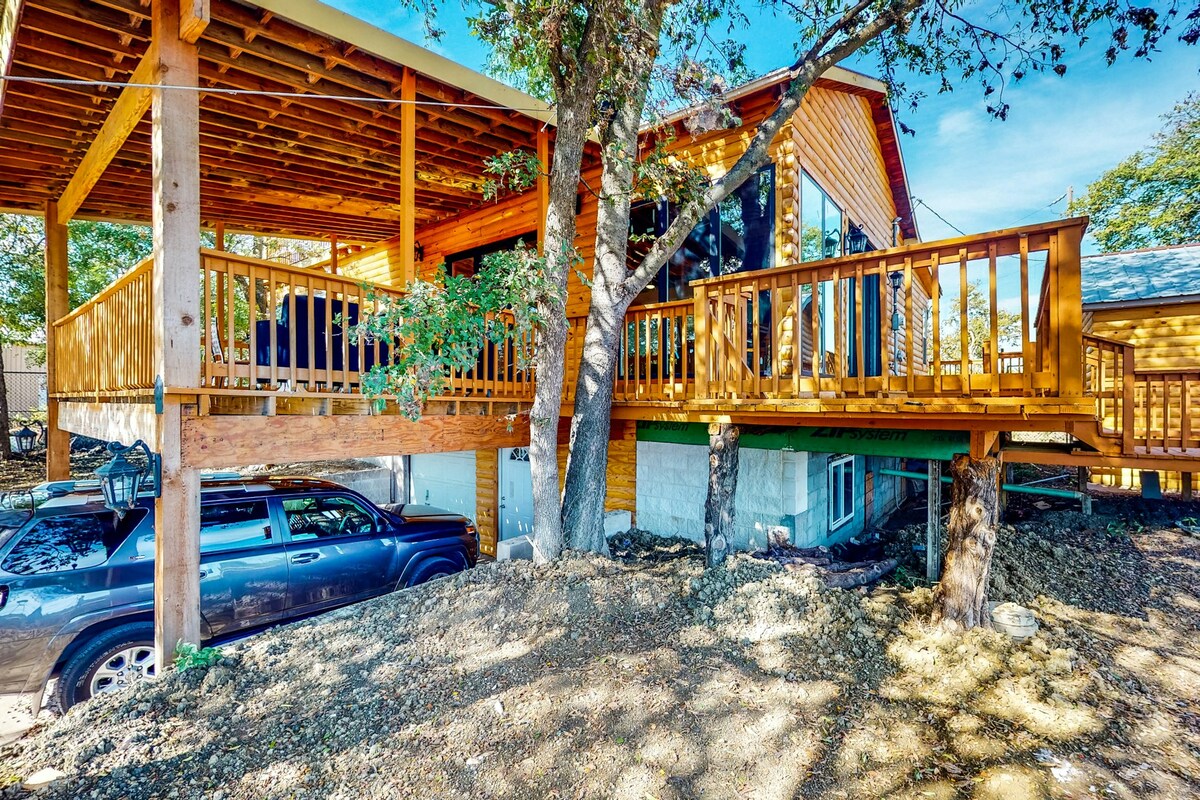 The exterior of the home features a multi-level design with a spacious deck elevated above the ground. Wooden railings surround the deck, while large windows reflect the surrounding trees. A vehicle is parked in the gravel area beneath the deck.