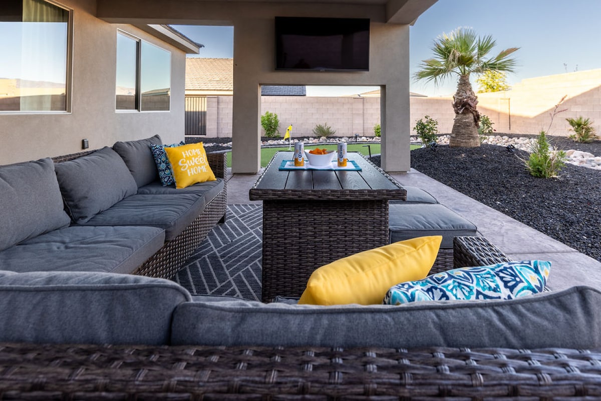 A shaded outdoor seating area features a large sectional couch with gray cushions and vibrant throw pillows, surrounded by a decorative coffee table. A wall-mounted TV is visible above, with desert landscaping and a palm tree seen in the background.