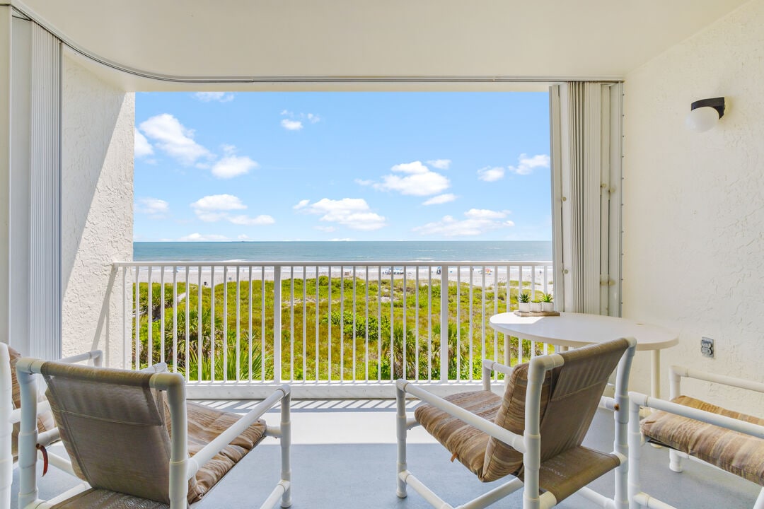 A balcony area features two comfortable chairs and a small table, offering a direct view of the ocean and sandy beach. Bright blue skies with fluffy clouds enhance the serene atmosphere, while green vegetation is visible in the foreground.