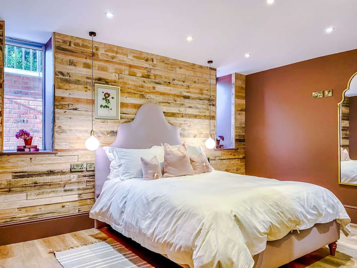 Genieße dieses warme und einladende Schlafzimmer mit einer rustikalen Holzakzentwand, die sanft von Pendelleuchten beleuchtet wird, mit einem Plüschbett mit Pastelltönen, das einen Hauch von Eleganz verleiht, und natürlichem Licht aus den Fenstern.