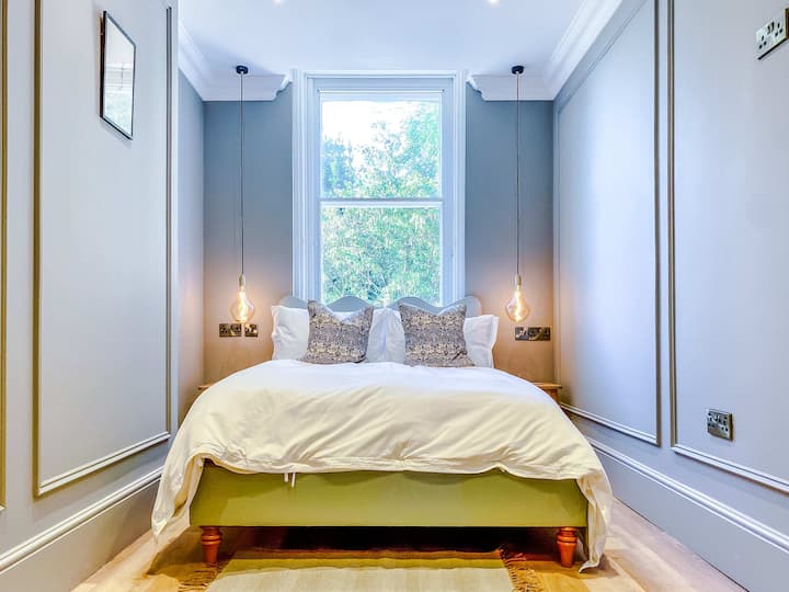 Ein ruhiges Schlafzimmer mit einem weichen, einladenden Bett, das mit frischer weißer Bettwäsche und Kissen bezogen ist, und einem großen Fenster, das einen Blick auf grüne Pflanzen bietet und ein ruhiges, raffiniertes Ambiente schafft, das perfekt für erholsamen Komfort ist.