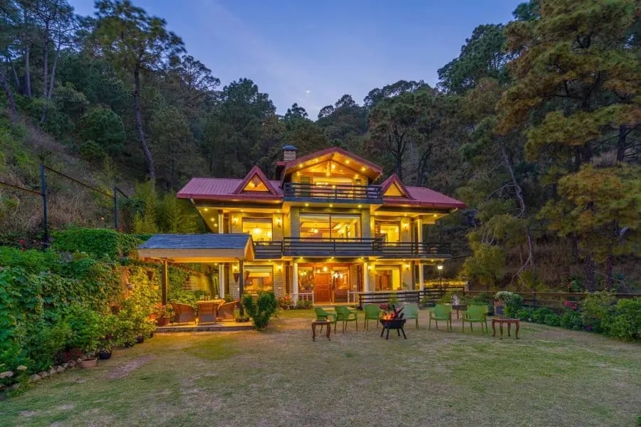 A three-story mountain chalet is surrounded by trees, illuminated warmly at dusk. The vibrant exterior is complemented by a spacious lawn featuring seating arrangements, a gazebo, and well-maintained greenery, creating an inviting outdoor setting for relaxation.