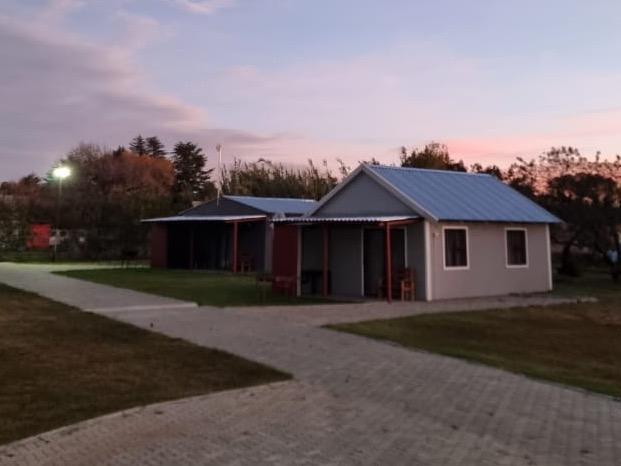 Two charming chalets are visible, each with a gabled roof and covered porch. A wide paved pathway connects them, surrounded by a well-maintained grass area. Soft evening light casts a warm glow over the scene, enhancing the tranquil setting.