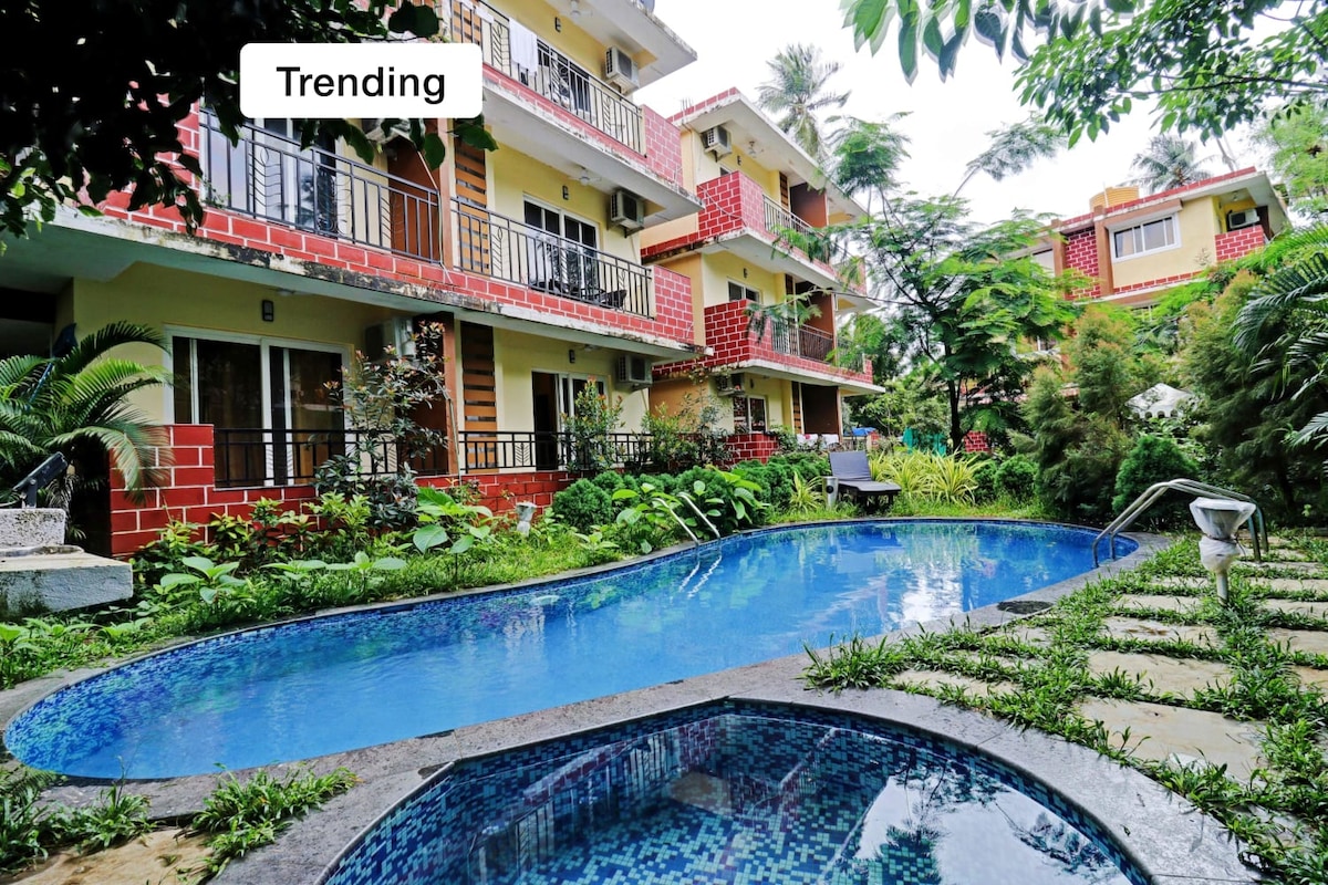 A tranquil outdoor space features a curved swimming pool, surrounded by lush greenery and tropical plants. Vibrant building facades with balconies are visible in the background, complementing the serene landscape.
