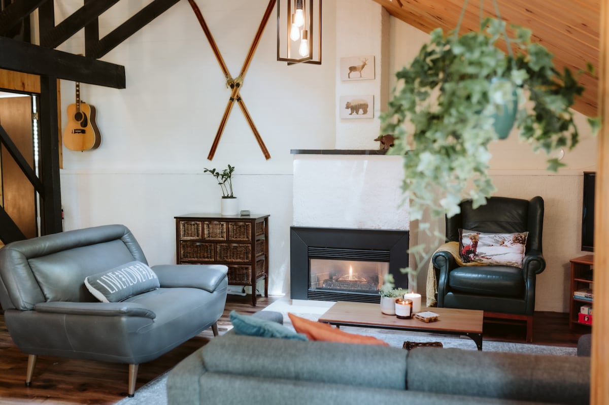 A cozy living area features a fireplace, surrounded by comfortable seating arrangements. Greenery hangs from the ceiling, complementing the rustic decor. Wooden beams and ski equipment adorn the walls, while natural light filters through nearby windows, creating a restful ambiance.