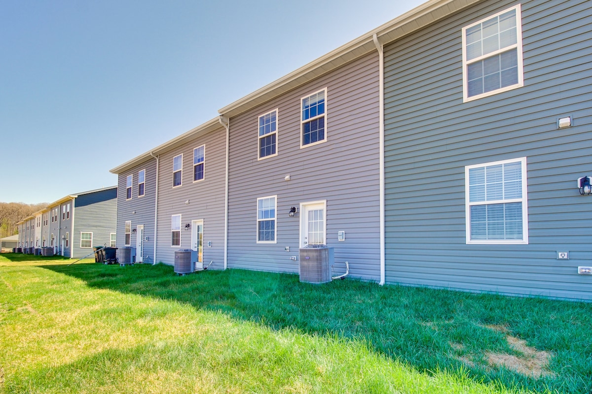A row of contemporary townhomes is showcased, featuring varied siding colors and multiple windows. Outdoor air conditioning units are visible, along with a grassy area that provides space for relaxation. The scene is brightened by clear blue skies.