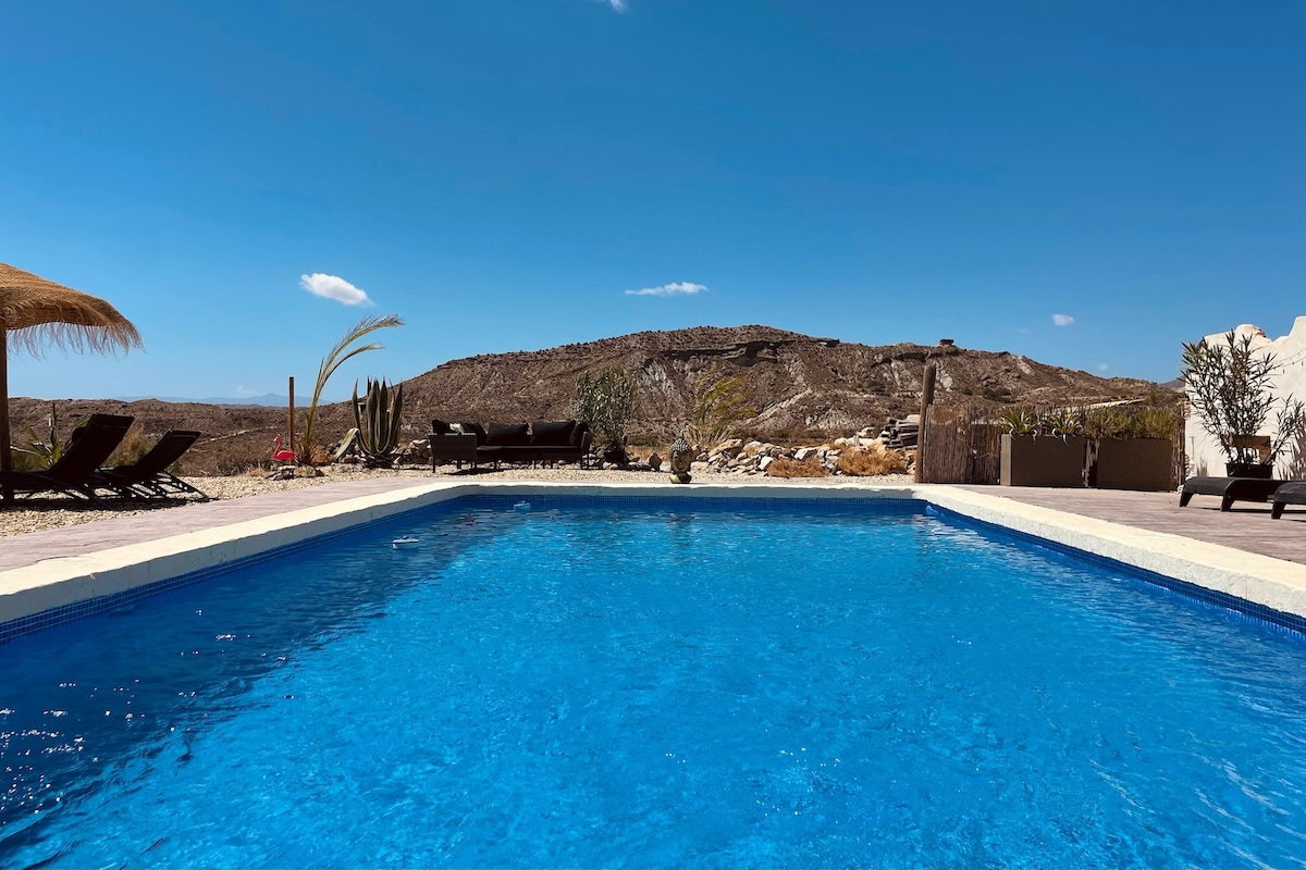 A refreshing swimming pool is featured at the property, surrounded by sun loungers. The landscape showcases arid terrain with distant hills under a clear blue sky. Cacti and sparse vegetation are visible near the pool area, contributing to the natural setting.