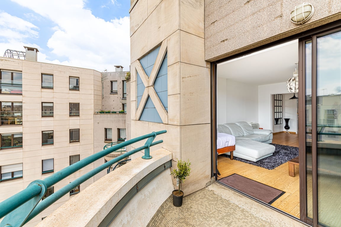 A spacious balcony is viewed from the apartment, featuring a comfortable lounge area and a soft rug. Light grey sofas are arranged for relaxation, with large sliding glass doors providing access to the indoor living space. The surface is tiled, and small greenery adds a touch of nature.