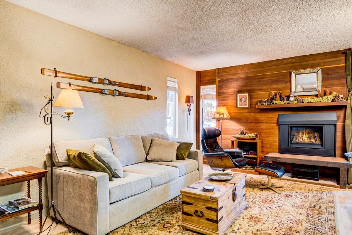 A bright living area is highlighted by a cozy gas fireplace with wooden accents and decorative elements. A soft, light-colored couch sits alongside a rustic coffee table, complemented by an accent chair. Gentle illumination is provided by lamps, enhancing the welcoming environment.