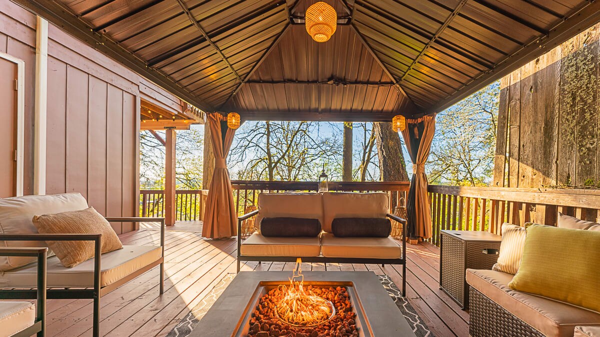 An outdoor space features a covered seating area with two cream-colored couches and a coffee table surrounding a fire pit. Warm lighting fixtures hang overhead, with large windows revealing views of surrounding trees and natural light streaming in.