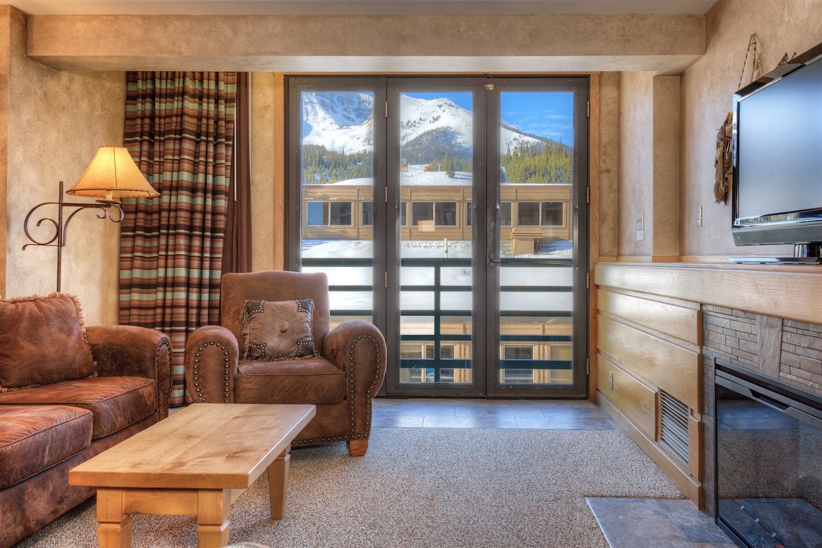 A comfortable living area features plush seating with a mix of leather and fabric furniture. A wooden coffee table is positioned at the center. Large windows provide stunning views of Lone Peak, while a flat-screen TV is mounted on the wall beside a stone fireplace.