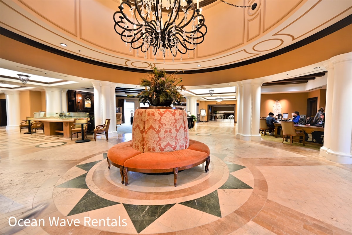 A spacious lobby area is highlighted by a circular seating arrangement with a plush orange upholstery. A decorative fountain with lush greenery sits at the center, while elegant chandeliers hang from the ceiling. Guests can be seen engaging in conversation, creating a welcoming environment.