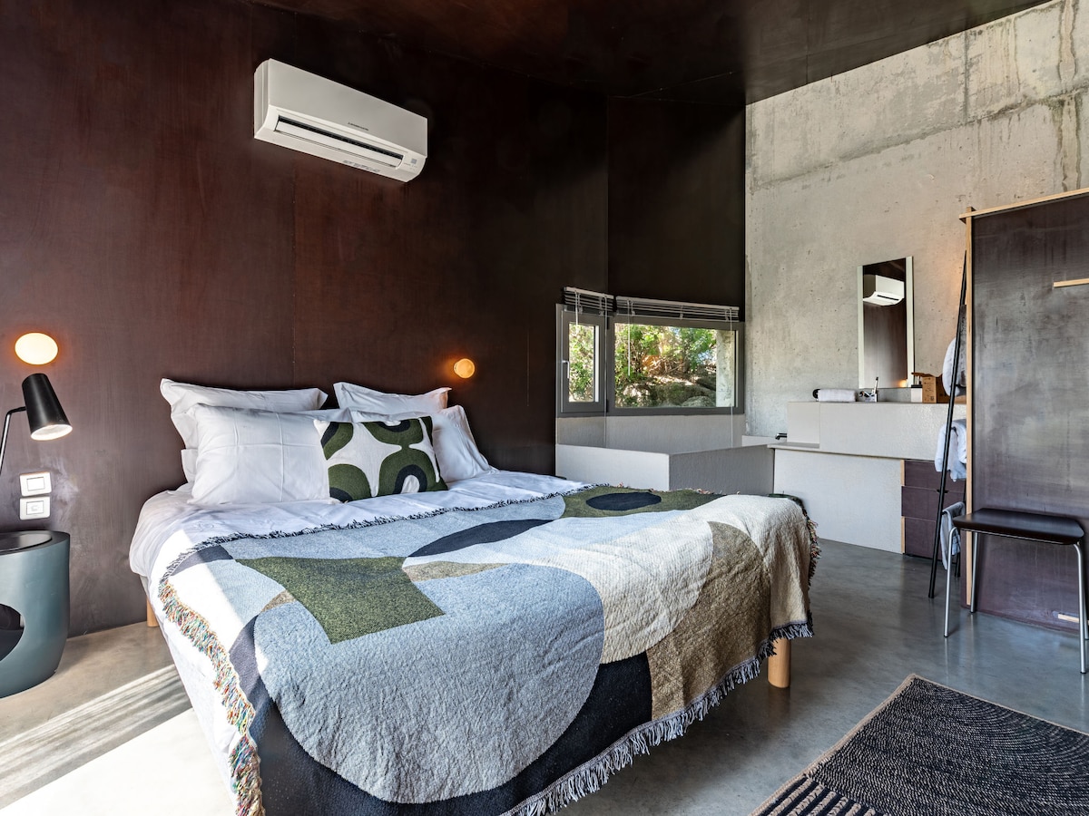 A well-appointed bedroom features a spacious bed adorned with a geometric throw and decorative pillows. Natural light streams from a window, illuminating the modern décor. Minimalist furniture, including a chair and a stylish bedside table, complements the concrete and wooden wall finishes.