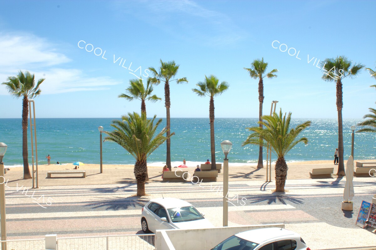 A view showcases the sandy beach and blue ocean, framed by palm trees. The coastline is bordered by a promenade with benches for relaxation, and distant figures can be seen enjoying the shoreline. The sky is bright with a few scattered clouds.