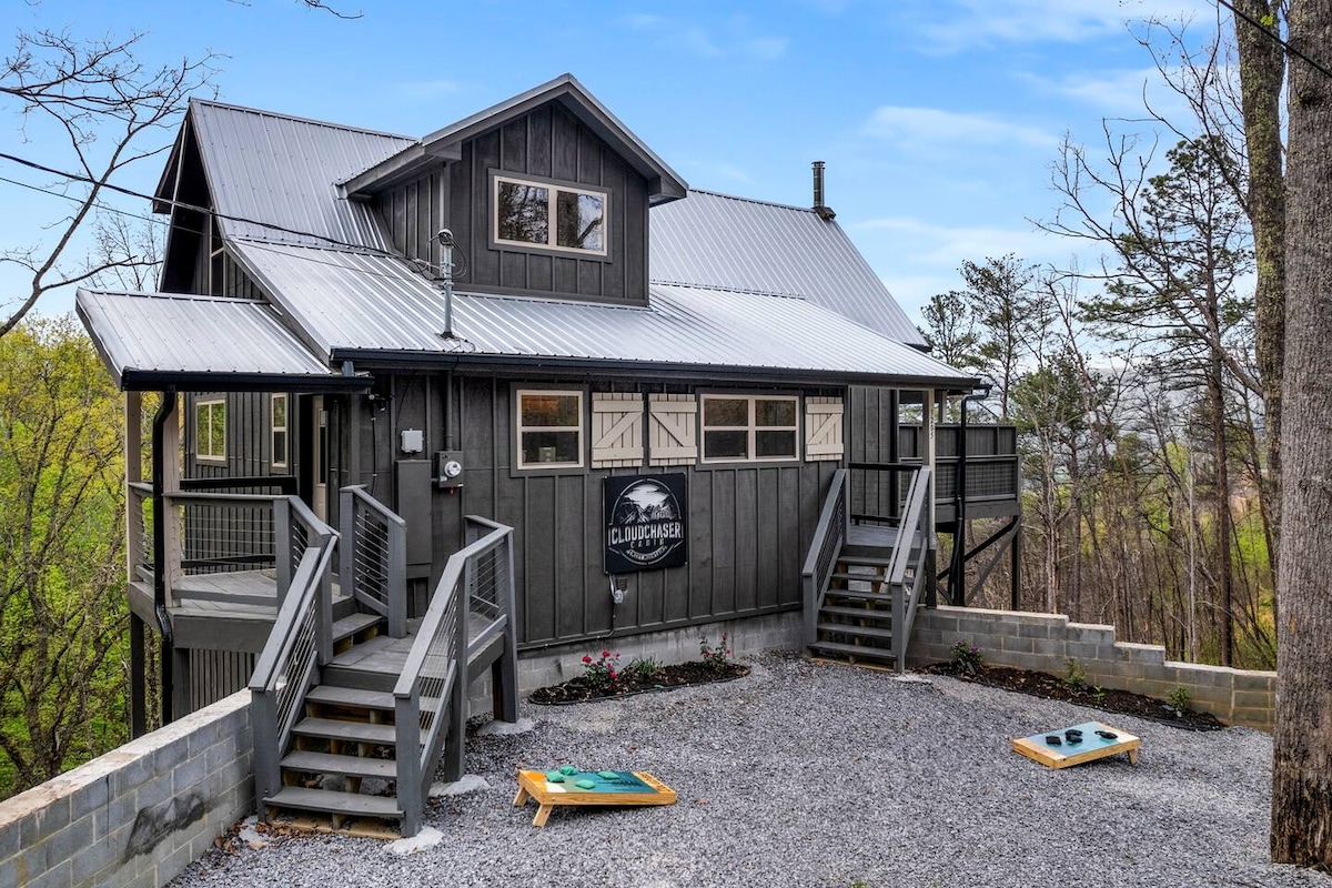 The exterior of Cloudchaser Cabin is presented, featuring a two-story structure with a metal roof and wooden siding. Sturdy steps lead to the entrance, and a gravel area with cornhole boards is visible, surrounded by trees and manicured landscaping.