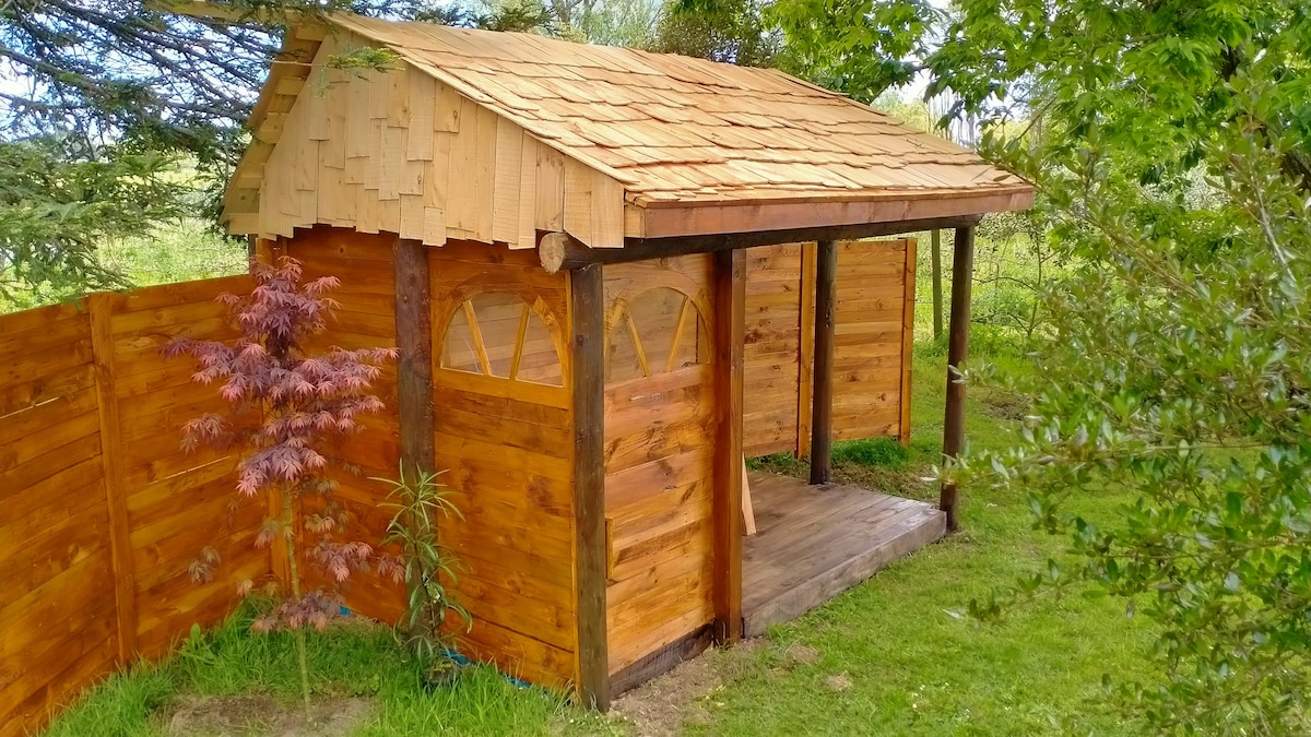 A rustic wooden structure is set amidst lush greenery, featuring a charming sloped roof with wooden shingles. The entrance is framed by wooden posts and complemented by a small plant nearby. Wooden fences enclose the space, offering a sense of privacy.