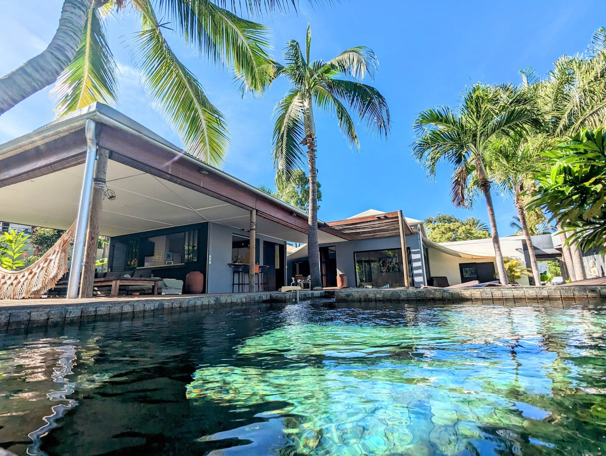 The image captures the inviting exterior of a villa, showcasing a shimmering pool surrounded by palm trees. A spacious terrace with seating is seen, blending with the tropical garden landscape. The villa's airy design offers large windows that provide views of the tranquil surroundings.