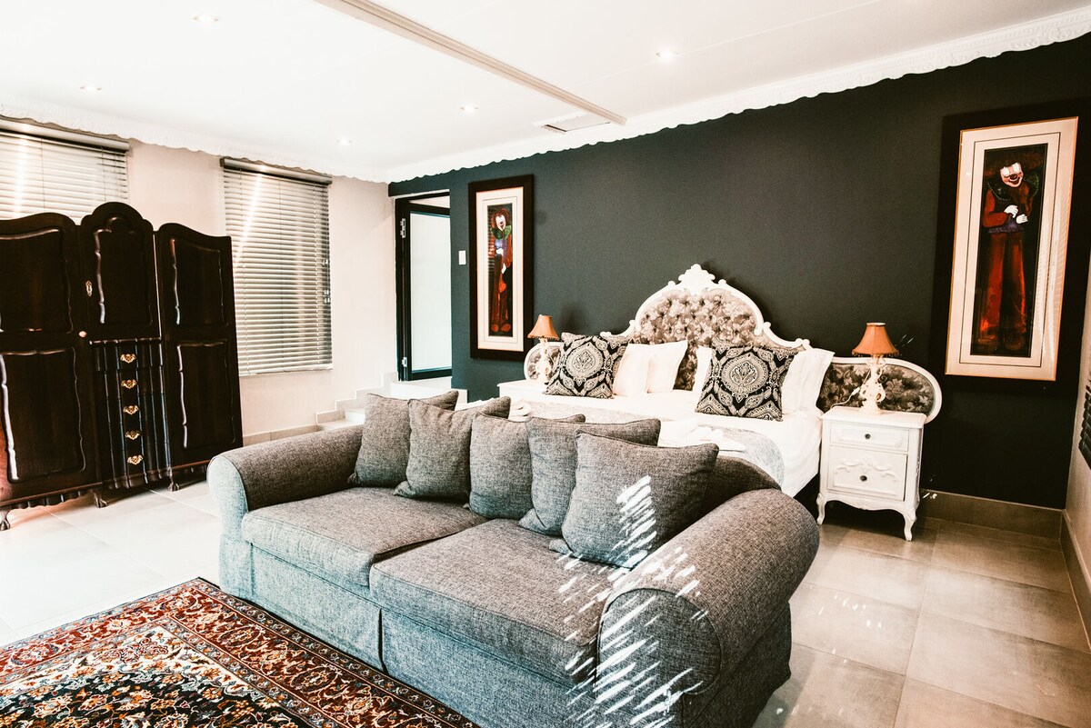 A spacious room features a large gray sectional sofa and a king-size bed, accented by decorative wall art. Soft lighting is provided by bedside lamps on either side of the bed, which is adorned with intricate patterns. Large windows allow natural light to fill the space.