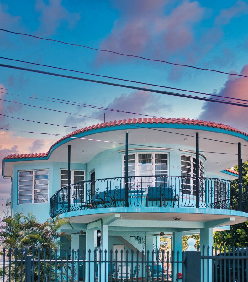 The house features a distinctive curved design with a vibrant blue exterior and a terracotta-tiled roof. A wrap-around balcony with black railings provides space for outdoor seating. Lush greenery is present in the foreground, framed by a gated entrance.