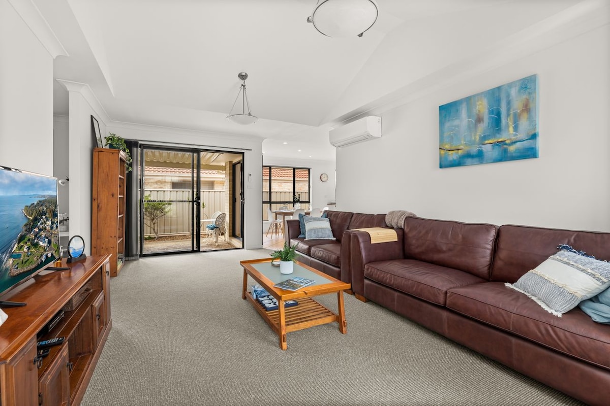 A spacious lounge area features a large brown leather sofa and a wooden coffee table. A flat-screen TV is positioned on a cabinet, while large sliding glass doors offer access to an outdoor space. Natural light brightens the room, complemented by neutral-toned walls.