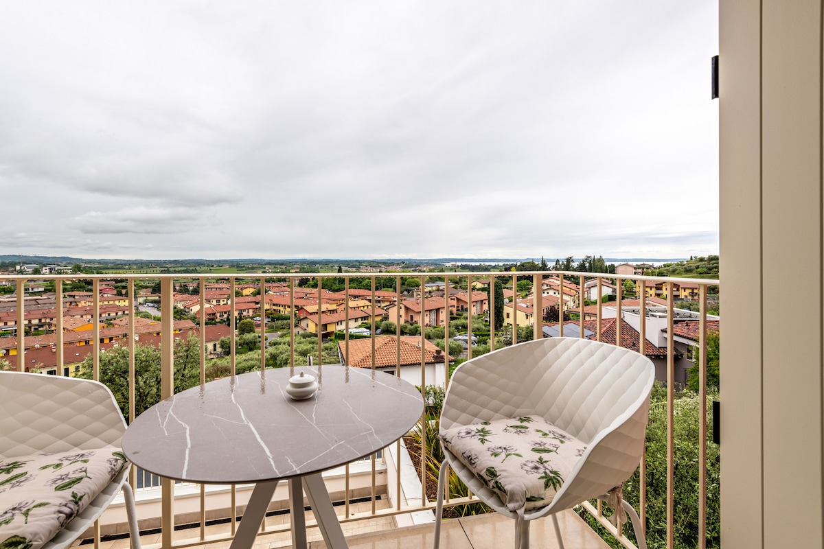 A private balcony is shown, featuring a circular marble table surrounded by two light-colored chairs. The view extends across the landscape, showcasing terracotta rooftops and lush greenery under an overcast sky.