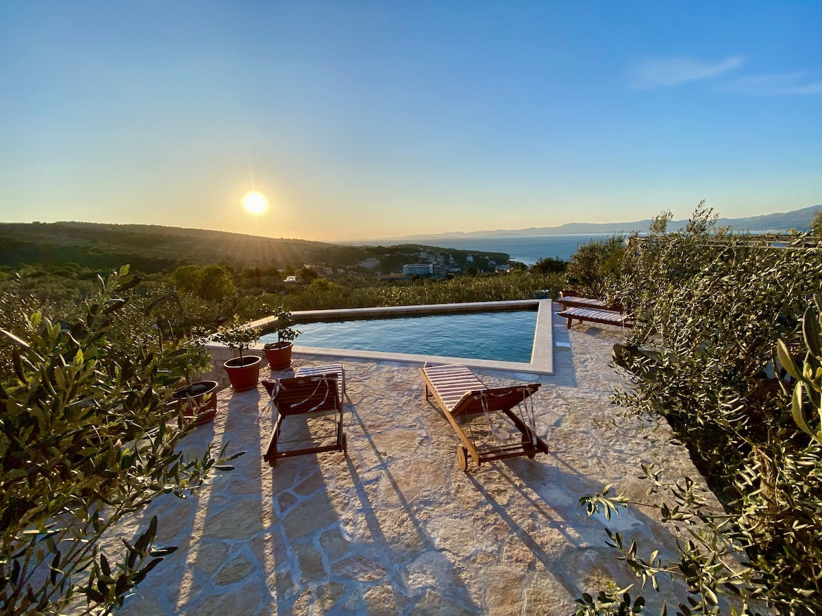 The image captures a serene view of a private swimming pool surrounded by olive trees, with lounge chairs positioned nearby. The sun sets in the background, casting a warm glow over the landscape, reflecting on the water's surface, with rolling hills and the sea visible in the distance.