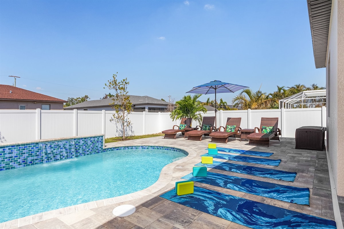 A tranquil backyard features a free heated pool surrounded by lounge chairs and umbrellas. Colorful pool floats rest on the water's edge, with vibrant tiles decorating the pool's interior. Lush greenery and a privacy fence create a serene atmosphere for relaxation.