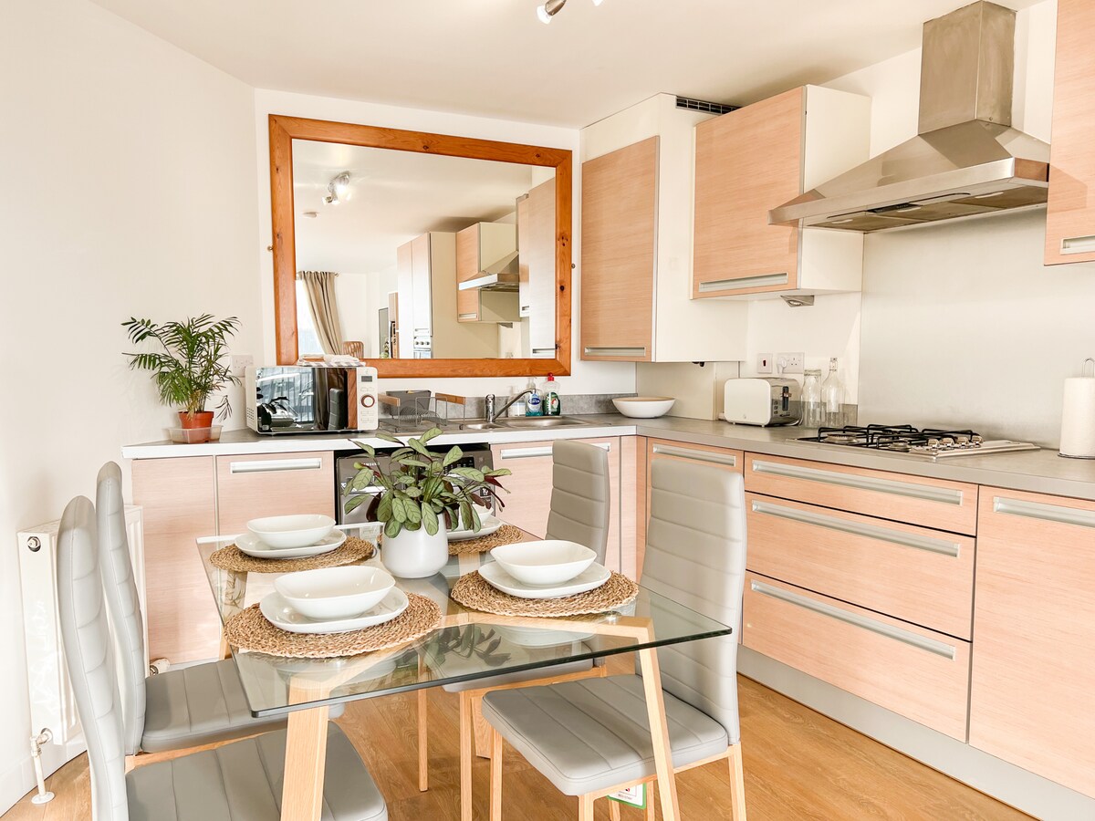 A modern kitchen area features light wood cabinetry and sleek appliances, including a microwave and gas stove. A glass dining table is set with four place settings surrounded by grey chairs, complemented by a potted plant on the table.