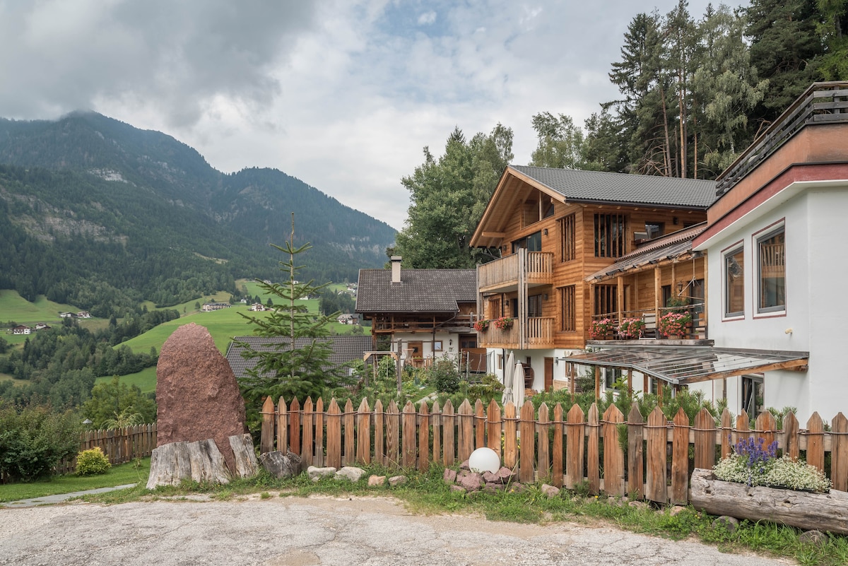 A charming building complex is framed by a wooden fence, with a large natural rock feature in the foreground. The two-story structures display wooden balconies and flower boxes, surrounded by lush greenery and mountains in the background, creating a serene rural atmosphere.