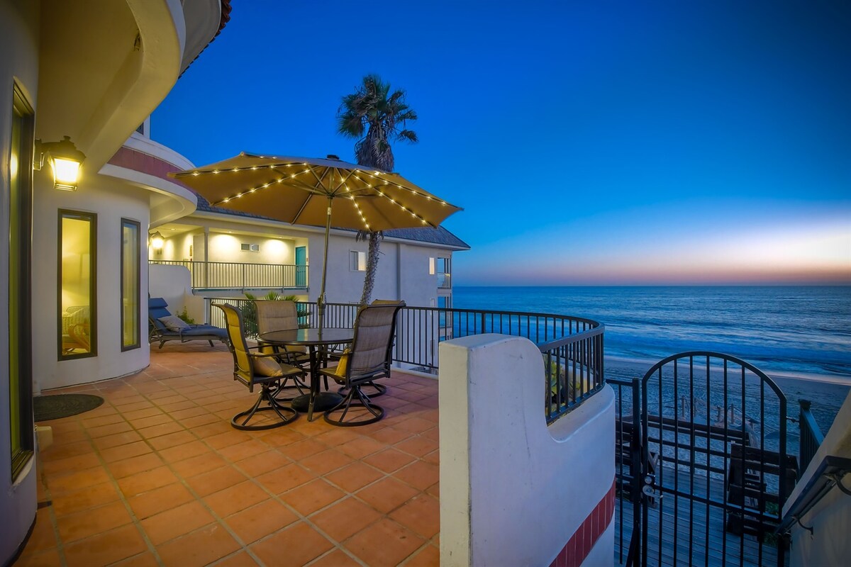 An outdoor patio area features a round dining table with seating for four, accompanied by an umbrella. Lounge chairs are positioned near the railing, which overlooks the ocean. The backdrop showcases a serene twilight sky, with soft hues transitioning into evening.