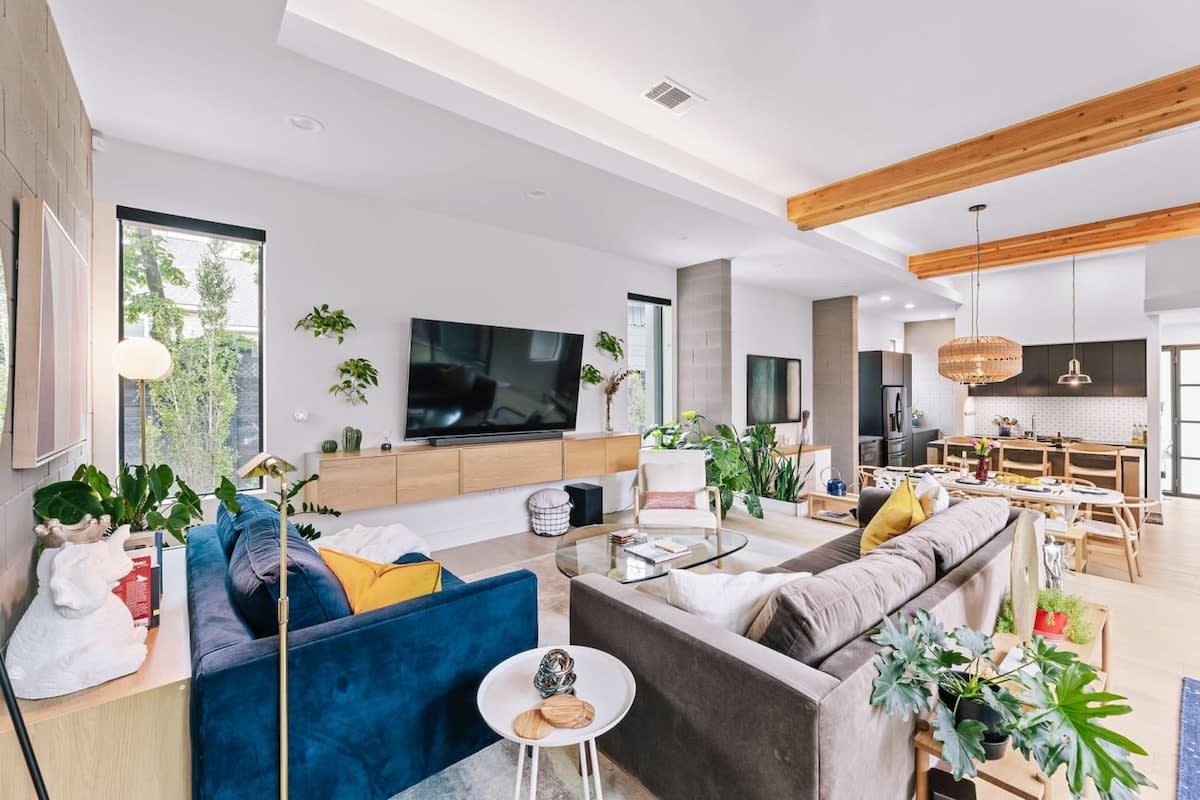 A spacious living area features comfortable seating arrangements with a mix of colorful cushions on two sofas. Large floor-to-ceiling windows allow natural light to flood the space, complemented by lush greenery and modern decor. A stylish dining area is visible in the background.