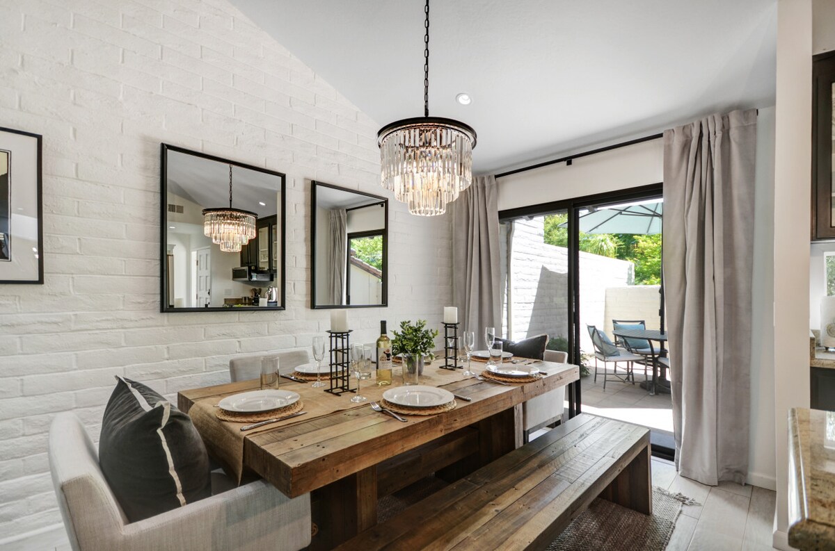 A rustic dining area features a wooden table set for six, surrounded by comfortable seating. Two mirrors reflect light and add depth, while a chandelier hangs above. Large sliding doors lead to an outdoor patio, enhancing the inviting ambiance.