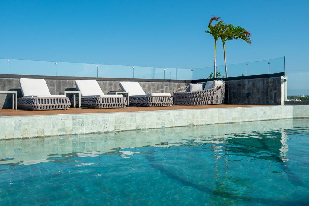A rooftop pool area features a shimmering water surface, bordered by elegant loungers and a woven seating arrangement. Lush palm trees sway gently in the background, complemented by clear blue skies and a modern glass railing.