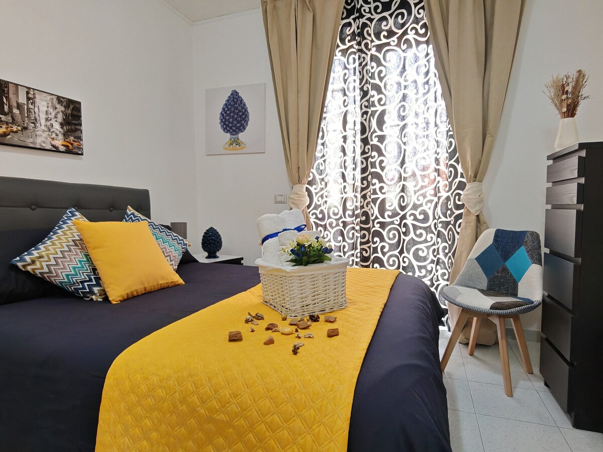 A well-appointed bedroom features a spacious bed adorned with a bright yellow throw blanket and cushions. A decorative chair sits nearby, and a small cabinet provides storage. Natural light filters through an elegant patterned window, illuminating the inviting space.