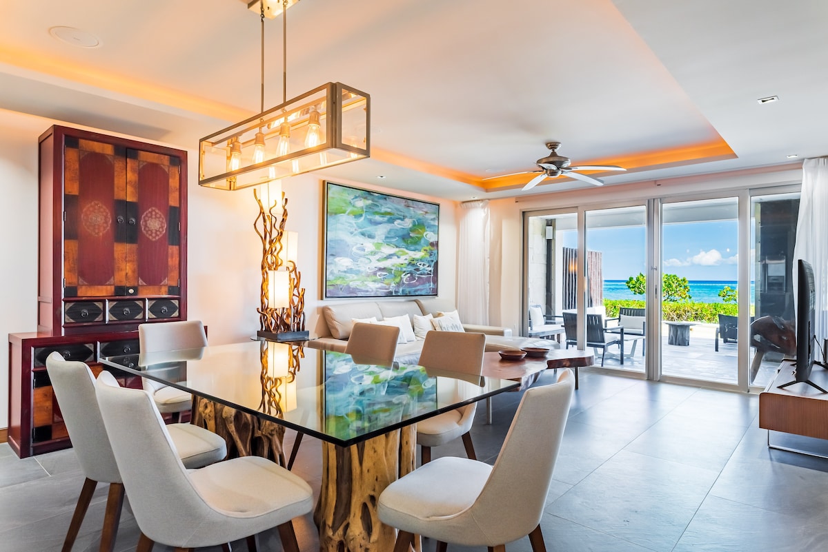 A spacious living area features a modern dining table surrounded by light-colored chairs. Large glass doors open to a patio with ocean views. Colorful artwork adorns the wall, complementing the natural wood accents and contemporary furnishings.