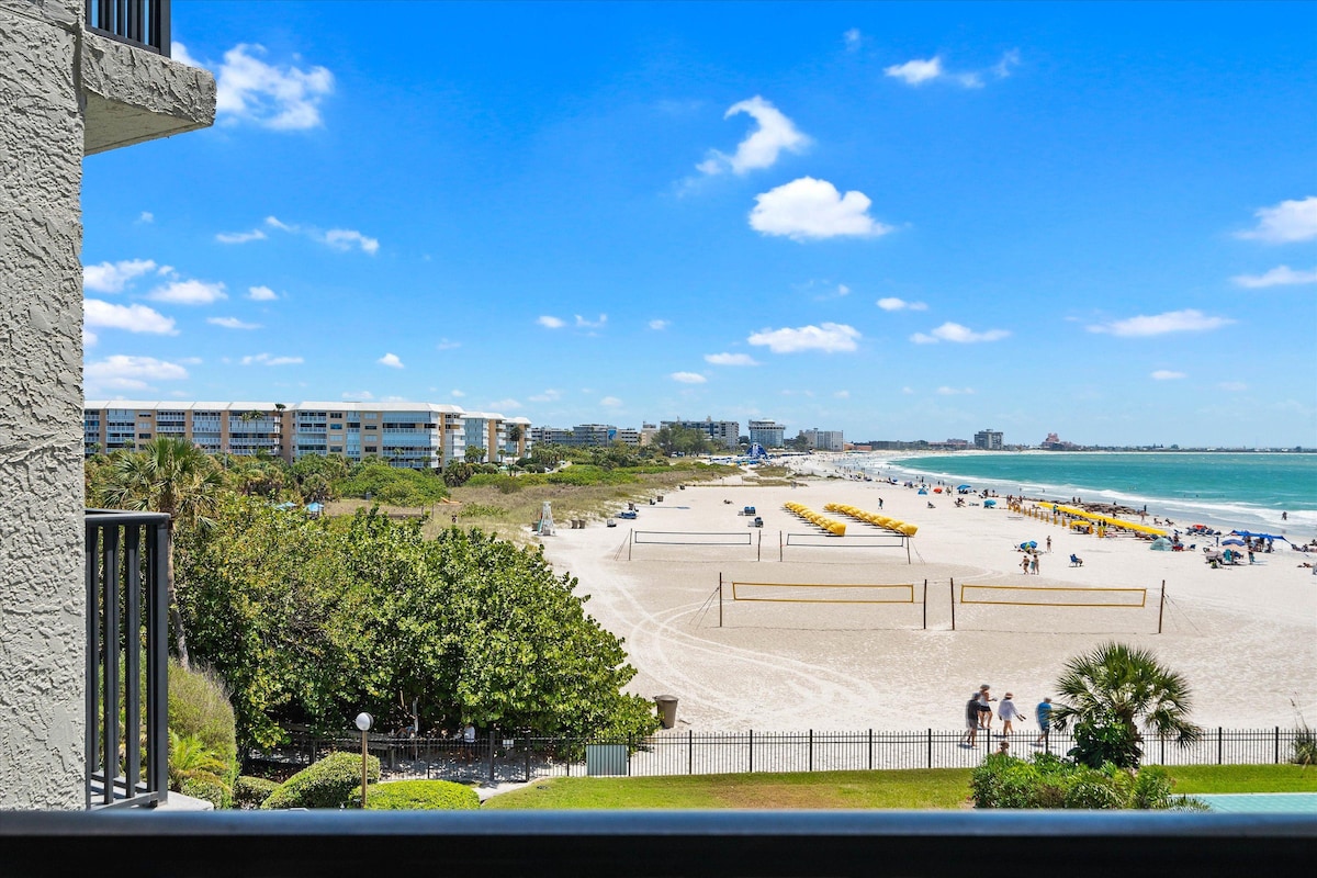 A panoramic view showcases Upham Beach and the Gulf of Mexico from the private balcony. Sandy beach areas are dotted with umbrellas and beachgoers enjoy the space. Lush greenery and volleyball courts create a vibrant coastal scene, framed by a clear blue sky.
