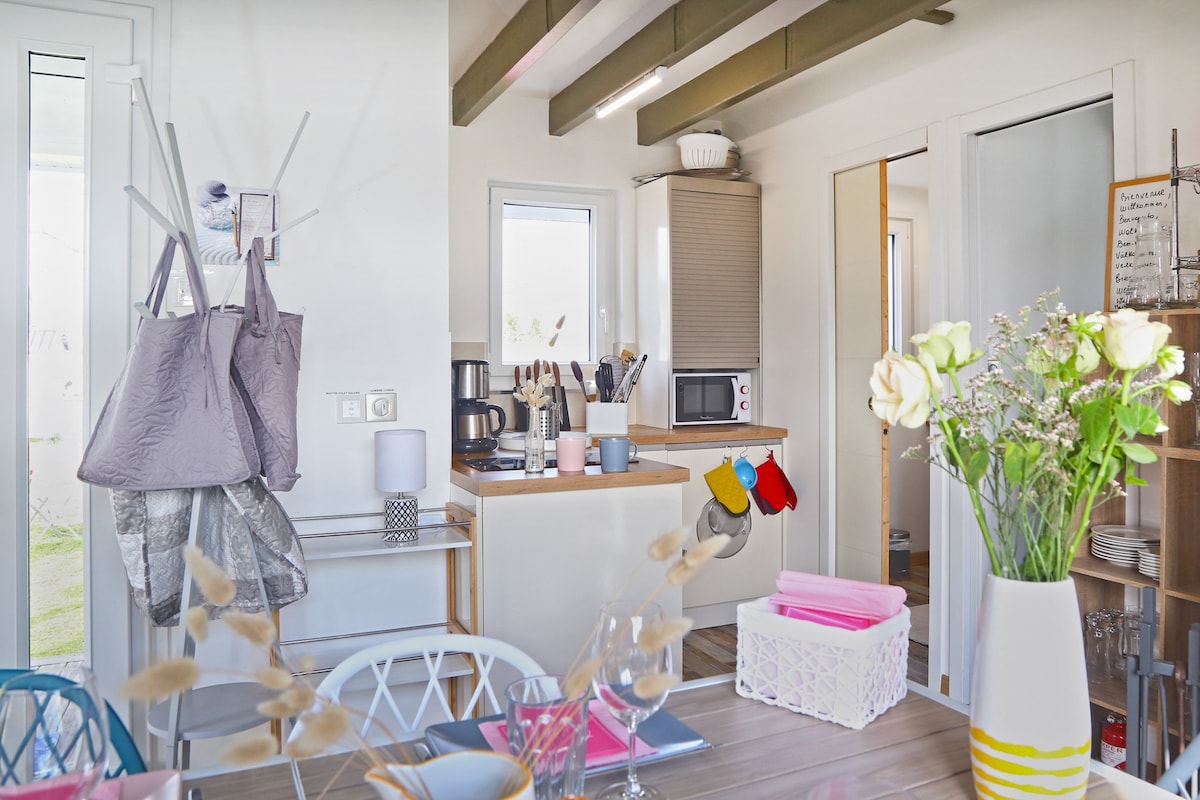 A functional kitchen area features a microwave, coffee maker, and oven, set against light-colored cabinetry. A dining table is adorned with pastel-coloured tableware. Hooks hold bags, and a vase with fresh flowers adds a touch of brightness. Natural light filters through the window.
