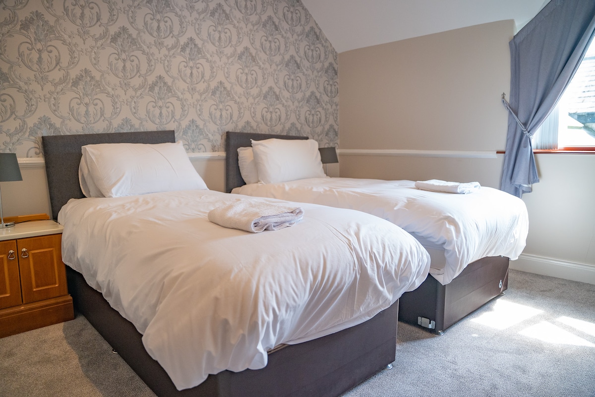 A well-lit bedroom features two spacious single beds with crisp white linens and neatly folded towels placed on top. The wall behind is adorned with subtle patterned wallpaper, and natural light enters through a partially draped window.