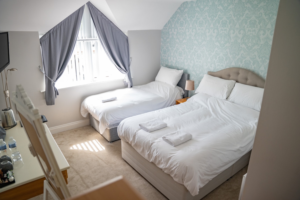 A bright guest room features two spacious beds, one double and one single, adorned with clean linens. Natural light enters through the window, which is dressed with soft grey curtains. A light blue patterned accent wall adds subtle color to the inviting space.