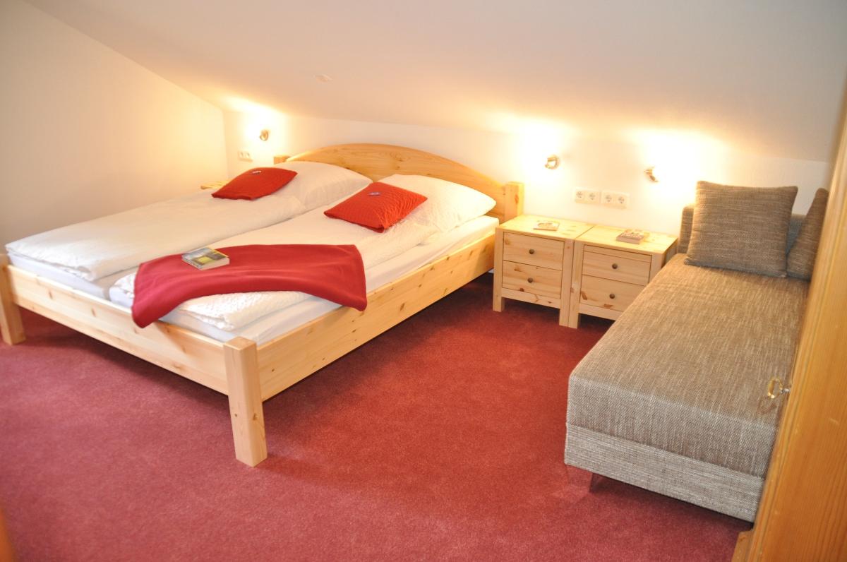 A serene bedroom features two separate beds topped with white bedding and red accent pillows. A cozy throw blanket rests on one bed. Two wooden nightstands with lamps are positioned beside the beds, while a comfortable lounge chair is placed against the wall.