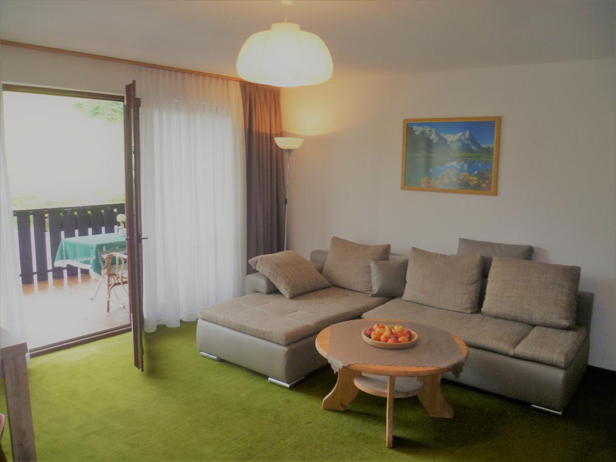 A comfortable seating area is highlighted by a large sectional sofa facing a round wooden table with fresh fruit. Natural light fills the space through an open door leading to a balcony, with a view of greenery beyond. A tasteful mountain artwork adorns the wall.