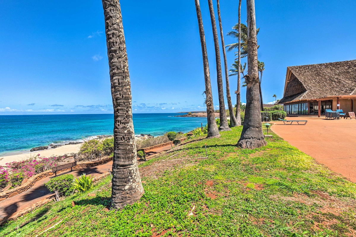 Palm trees frame a panoramic view of the ocean and a sandy beach. The property features a spacious patio area with lounge chairs, while vibrant foliage adds a touch of color to the landscape, enhancing the natural coastal setting.