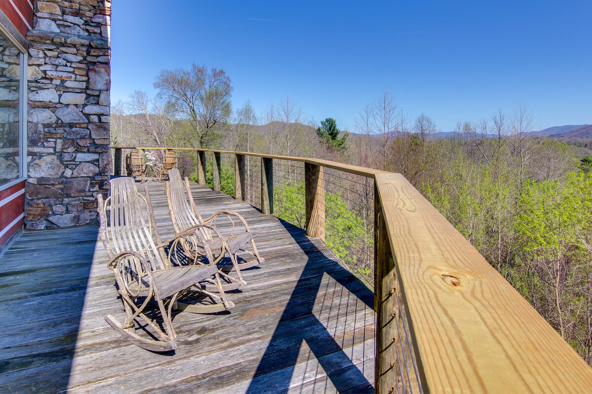 A spacious wraparound deck features multiple rocking chairs, offering panoramic views of the surrounding trees and rolling hills. The wooden railing highlights the natural surroundings, and the bright sky adds to the serene outdoor atmosphere.