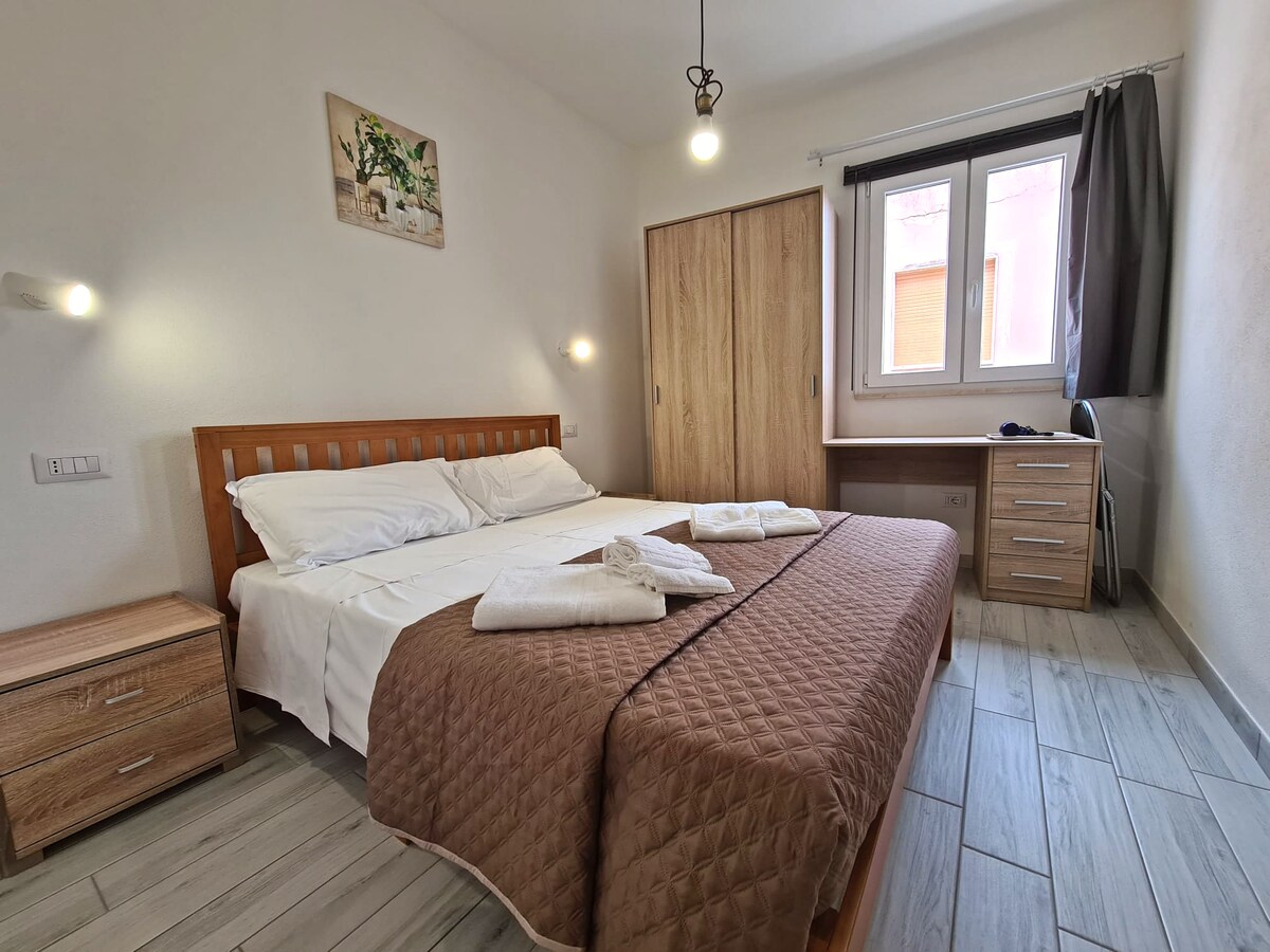 A cozy bedroom features a spacious wooden bed with a neutral-tone quilt. Two windows allow natural light to illuminate the space. A wooden desk and chair are situated near the window, while a bedside table holds an additional light source.